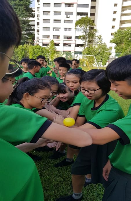 Students in green shirts huddle together on a grass field, balancing a small yellow ball on their joined wrists.